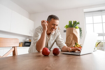 Mature man with apple using laptop at table in kitchen