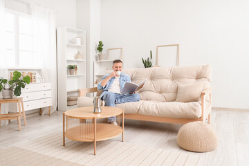 Mature man with cup of hot coffee reading magazine on sofa at home