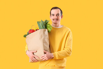 Young man with paper bag of fresh vegetables on yellow background