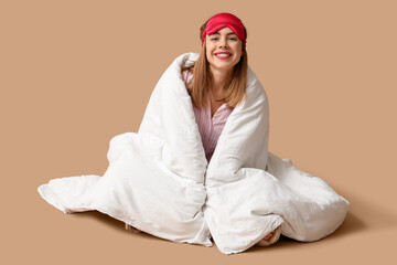 Beautiful young happy woman with soft blanket and sleeping mask sitting on brown background