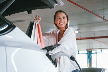 Young woman travel with EV electric car to shopping center parking lot charging in downtown city...