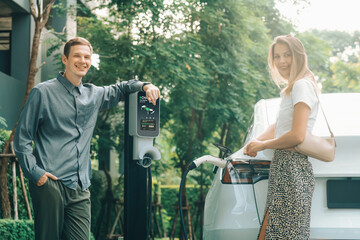 Young couple travel with EV electric car charging in green sustainable city outdoor garden in...