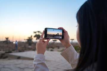 cute young woman holding a cell phone shooting the sunset scene from luxor temple, Luxor, Egypt.tourist exploring luxor templr at sunset. - obrazy, fototapety, plakaty