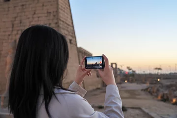 Foto op Plexiglas cute young woman holding a cell phone shooting the sunset scene from luxor temple, Luxor, Egypt.tourist exploring luxor templr at sunset. © Mostafa Eissa