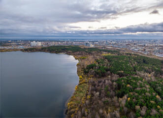 Autumn forest on lake shore at sunset and city on horizon, aerial view