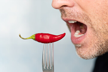 Expressive and funny man holding whole red chili pepper in his mouth. Man eating a spicy pepper on...