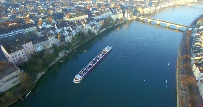Stunning sunrise captured by a drone: a cargo ship in the Rhine river, leaving behind the bridge after emerging from it, moving forward in the morning light of Basel, Switzerland.