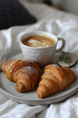 A cozy breakfast scene with a cup of coffee with latte art and two fresh croissants on a ceramic plate, adorned with a sprig of greenery.