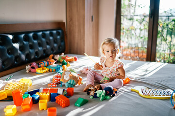 Little girl sits with a wand in her hand near a xylophone on a bed surrounded by soft toys