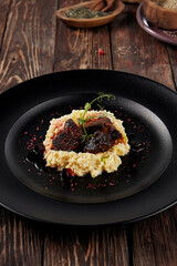 Braised beef cheeks with millet on a black plate, served on a rustic wooden table