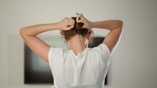 Woman ties hair into ponytail in home gym. preparing for jogging. Dynamic athlete dressed in sports top arranges short hair running on treadmill indoors