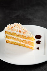 A slice of white chocolate and coconut cake on a white plate with dark syrup, against a textured black backdrop