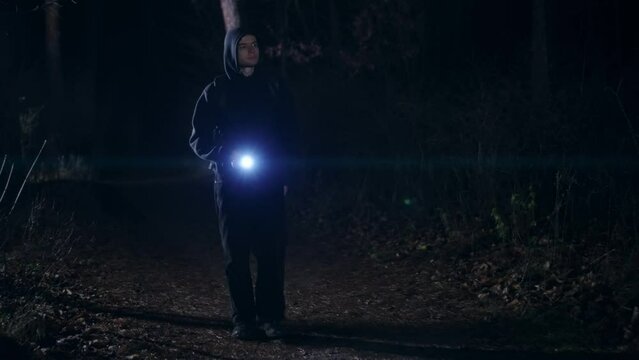 A young guy with a backpack on his back walks through a forest or park at night. A man in a hoodie and with a flashlight in his hand