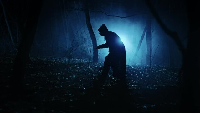 Mysterious dark figure in the night foggy forest. Silhouette of an unrecognizable man in a tunic with a hood.