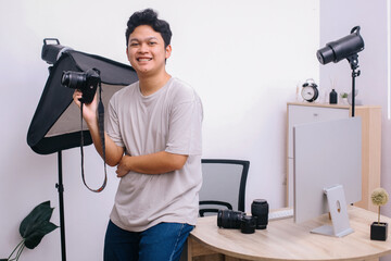 Portrait of young Asian photographer smiling while holding DSLR camera at photo and video production studio office.
