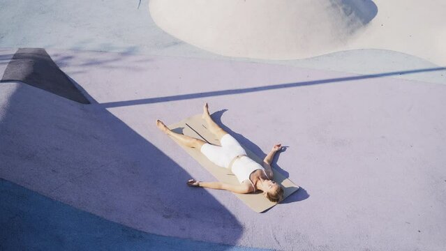 Young woman in white tracksuit lying down in deep relaxation during yoga training at urban sports ground in sunny morning. Camero motion shot, Lady practicing Dead Body Corpse Pose ,Shavasana