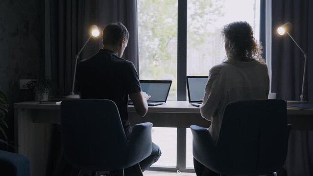Couple of freelancer silhouettes working opposite bright window with contemporary laptops. Back side view unrecognizable man and woman kissing while working at home