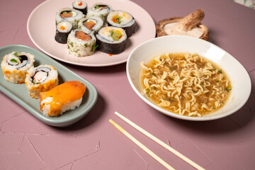 Chop sticks, mushrooms, noodle ramen, California rolls and Philadelphia rolls on a small plate on a pink background
