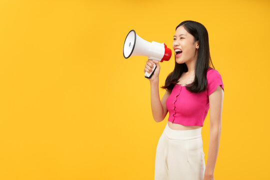 Portrait of a young Asian woman holding a megaphone and shouting aggressively. Wearing a pink t-shirt, and isolated on a yellow background.