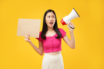 Portrait of an excited Asian lady holding a megaphone and paper, looking at the camera and isolated on a yellow background. 