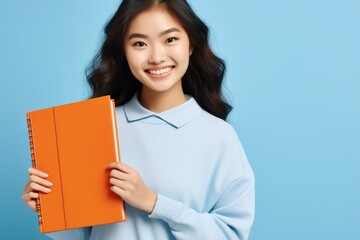 Obraz premium Young Asian woman smiling, holding orange notebooks against a blue background.