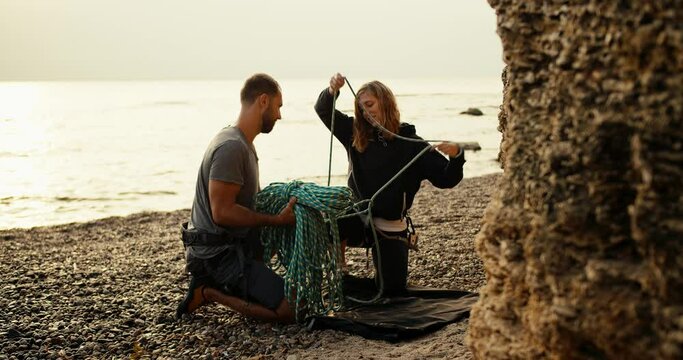 A blonde girl with curly hair in black clothes untangles and unties a special rope together with her boyfriend in order to start rock climbing on a rocky shore near the rocks