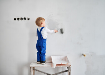 Back view of child construction worker standing on wooden table and painting white wall in apartment. Kid in work overalls using paint roller while working on home renovation.