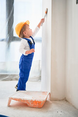 Full length of child construction worker painting wall with paint brush in living room. Kid in...