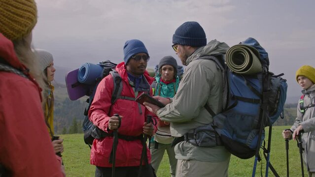 Multi ethnic travelers discuss trail way using tablet computer on mountain hill. Hiking buddies with backpacks and trekking poles stop to rest during expedition. Concept of tourism and active leisure.