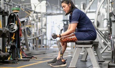 Woman with prosthetic leg sitting in gym lifting dumbbell weight. Female with foot prosthesis...