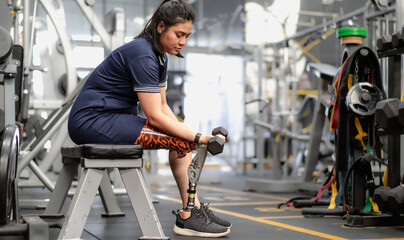 Fototapeta na wymiar Woman with prosthetic leg sitting in gym lifting dumbbell weight. Female with foot prosthesis physical workout exercise in fitness. Artificial limb equipment help accident survivor amputee to mobility