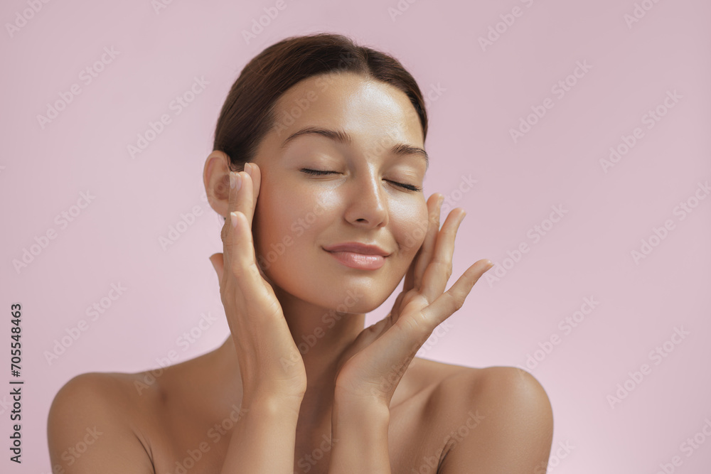 Wall mural Skincare and Beauty Portrait. Woman with beautiful face touching healthy facial skin. Young adult smiling Asian girl model with natural makeup touching glowing hydrated skin on pink background closeup