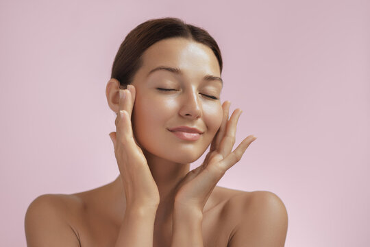 Skincare and Beauty Portrait. Woman with beautiful face touching healthy facial skin. Young adult smiling Asian girl model with natural makeup touching glowing hydrated skin on pink background closeup