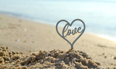 Plastic stick in shape of heart and word Love in sand on beach seashore on sunny summer day close-up. Figures in shape of heart word Love on background of sea waves. Love relationship romance concept - obrazy, fototapety, plakaty
