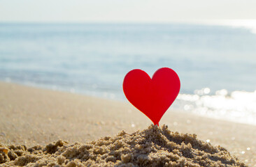 Plastic stick in shape of red heart in sand on sandy beach of sea shore on background of sea waves...