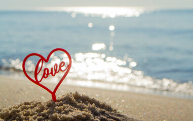 Fototapeta premium Plastic stick in shape of red heart and word Love in sand on beach seashore on sunny summer day close-up. Figures in shape of heart word Love on background sea waves. Love relationship romance concept