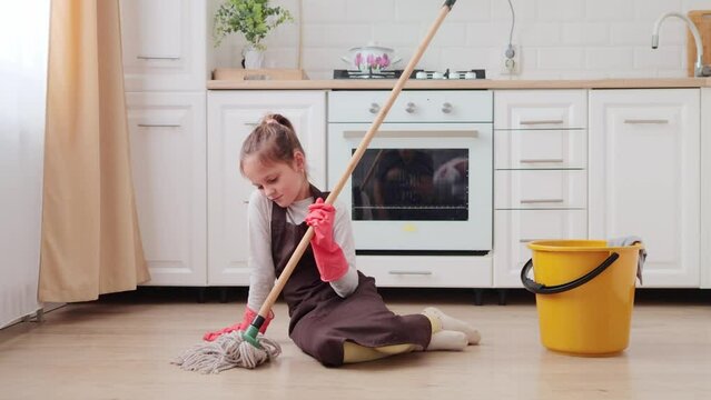 Sad upset bored little girl sitting in kitchen and washing floor with mop wants playing with friends instead of cleaning the house kid wearing apron and rubber gloves