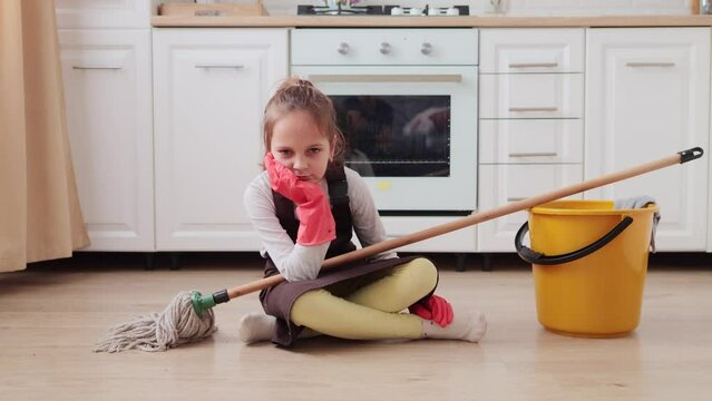 Bored unhappy little female kid in apron and gloves sitting on floor holding mop in hands looks sad and exhausted being tired to do lots work about the house cleaning apartment