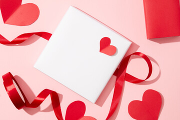 A white card, red ribbon and red paper hearts on a pastel pink background. Empty space for text design. Copy space viewed from above. Love theme.