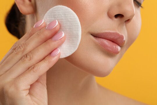 Beautiful woman removing makeup with cotton pad on orange background, closeup