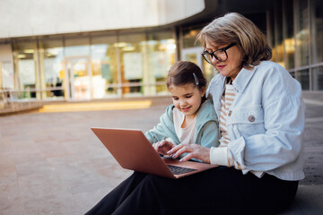 A middle-aged woman and a little girl are sitting outdoors with a laptop.