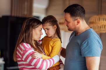 Young parents comfort their cute daughter. Beautiful woman hugs and talks to little girl in living...