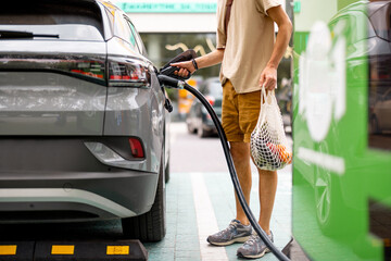 Man with groceries waiting for electric car to be charged on public charging station near a...
