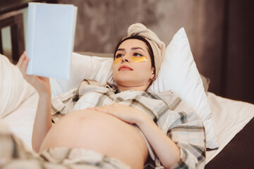 Young happy pregnant woman in pajama reading book and touching belly gently while relaxing at home.