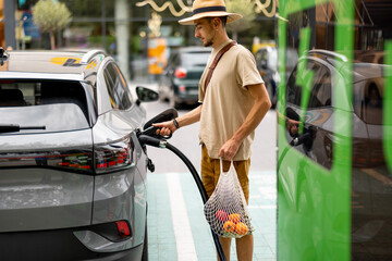 Man with groceries waiting for electric car to be charged on public charging station near a...