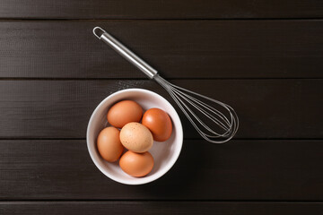 Metal whisk and raw eggs in bowl on dark wooden table, top view
