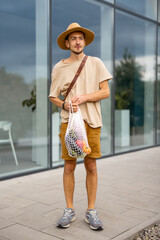 Stylish man in hat walks with a mesh bag full of fresh vegetables near supermarket outdoors. Concept of sustainable lifestyle and style