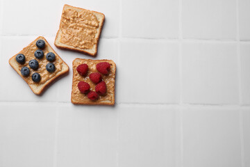 Delicious toasts with peanut butter, raspberries and blueberries on white tiled table, flat lay. Space for text