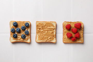 Delicious toasts with peanut butter, raspberries and blueberries on white tiled table, flat lay