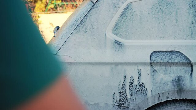 Washing a car with shampoo and foam under high pressure at the dacha in sunny weather. Contactless shampoo for cleaning dirt from the body of a gray car. Slow motion